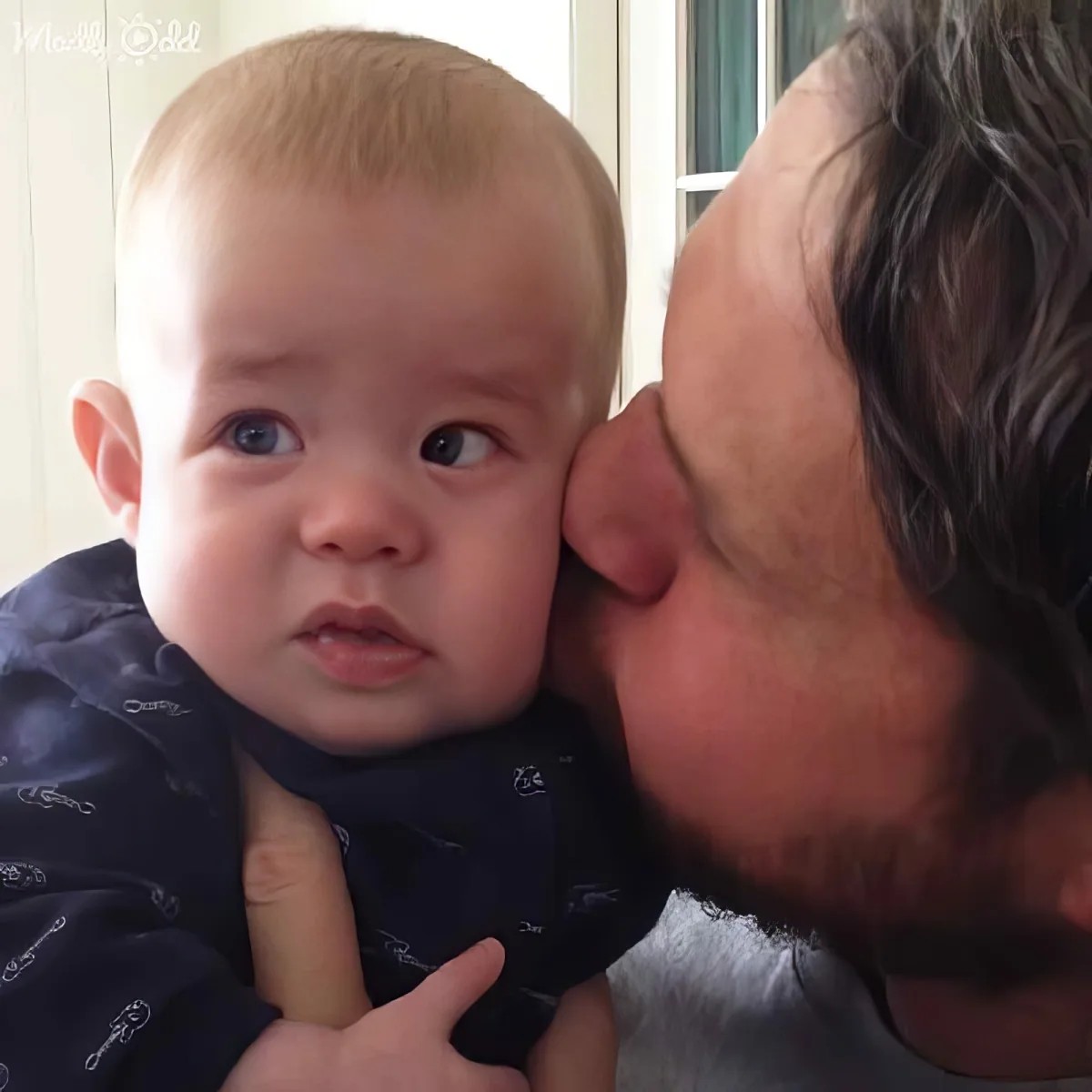 A man with a beard holds a baby, looking at the camera with a curious expression.
