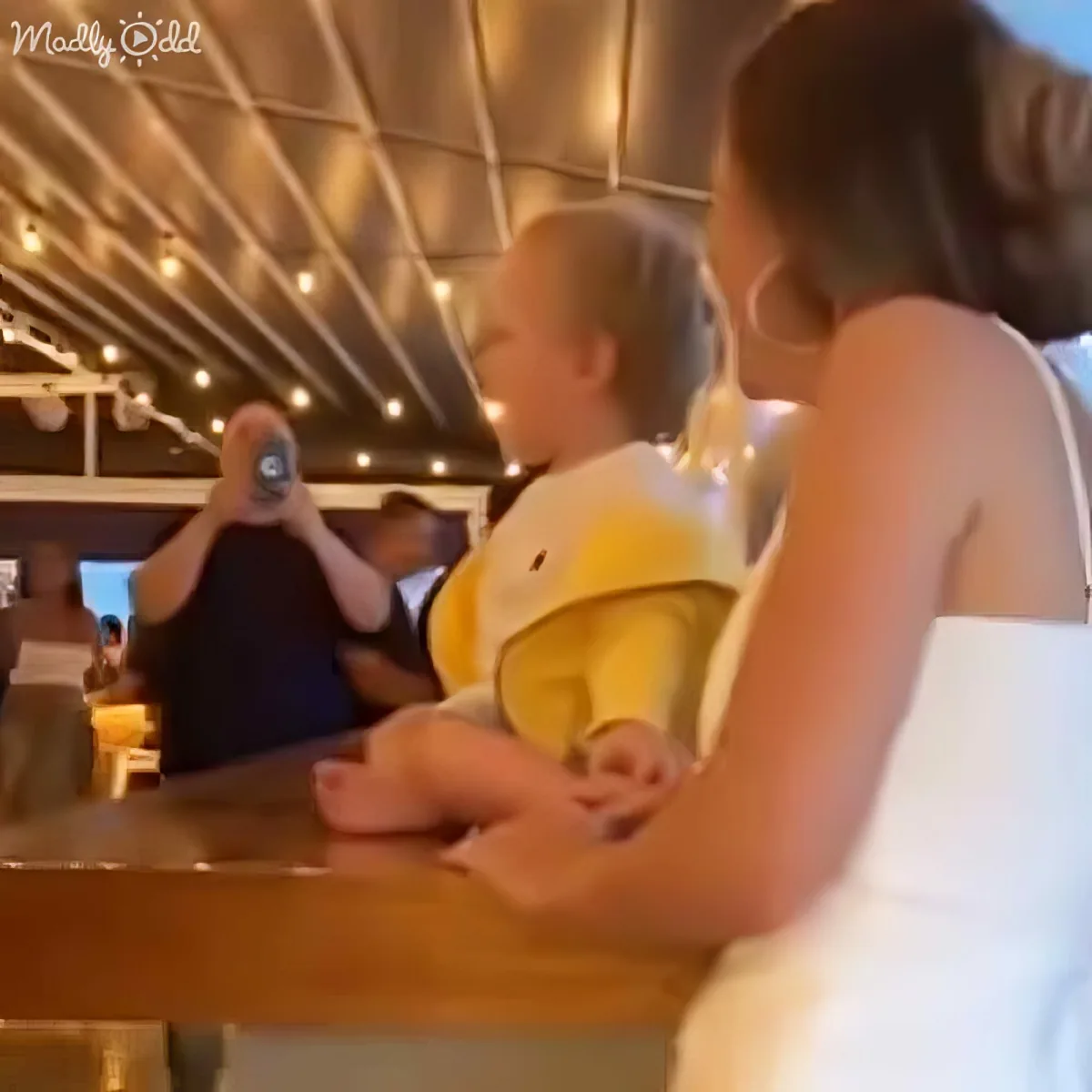 A baby in a yellow outfit is being held by a person at a bar, with a ceiling of lights and other people in the background.