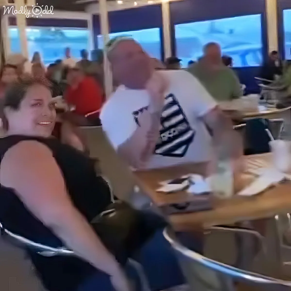 A man in a white shirt is playfully leaning over a table, interacting with a woman in a black top at a restaurant.