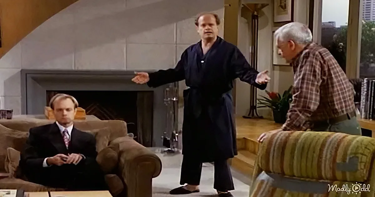 A man in a robe gestures dramatically while another sits in a suit, and a third leans on a striped couch in a living room.