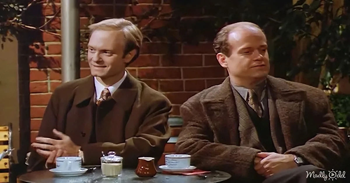 Two men in brown suits sit at a table with coffee cups, smiling and looking to the side.