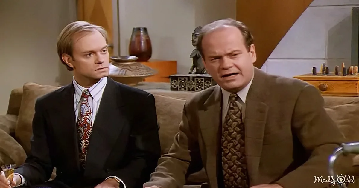 Two men in suits sit on a couch, one looking concerned, the other with a surprised expression.