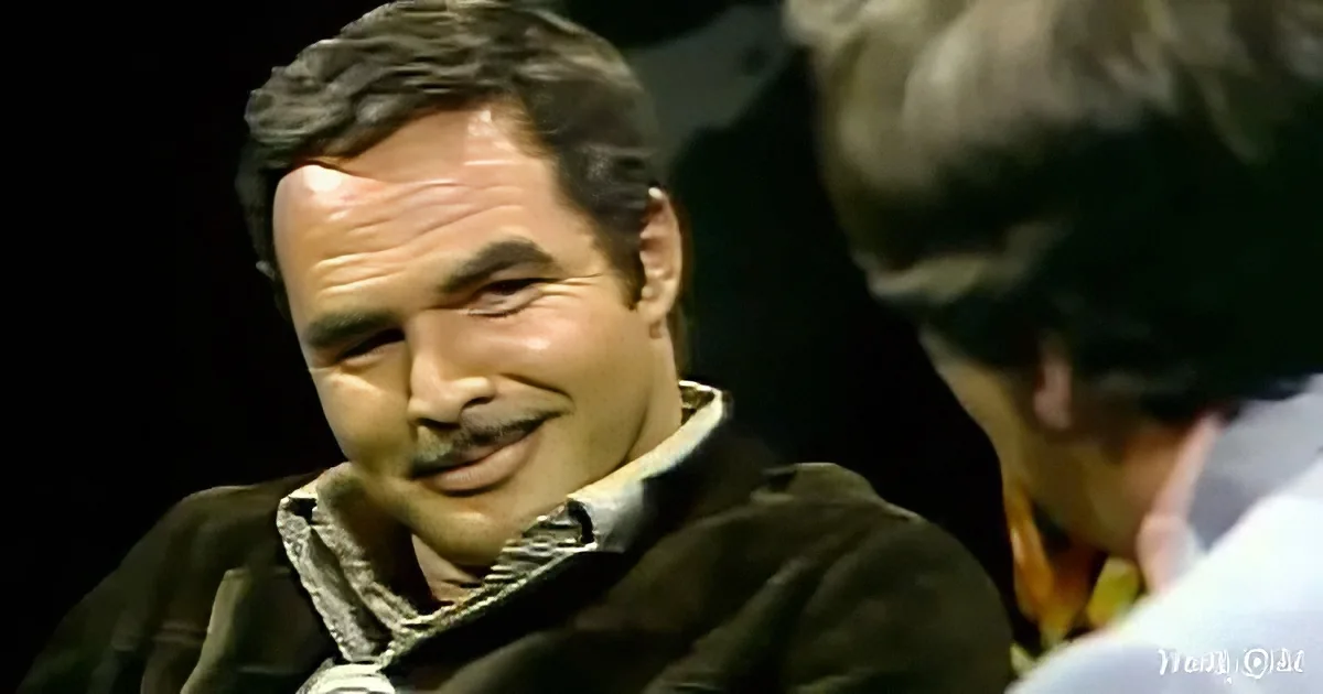 A man with a mustache and a dark jacket smiles while another person is partially visible behind him.