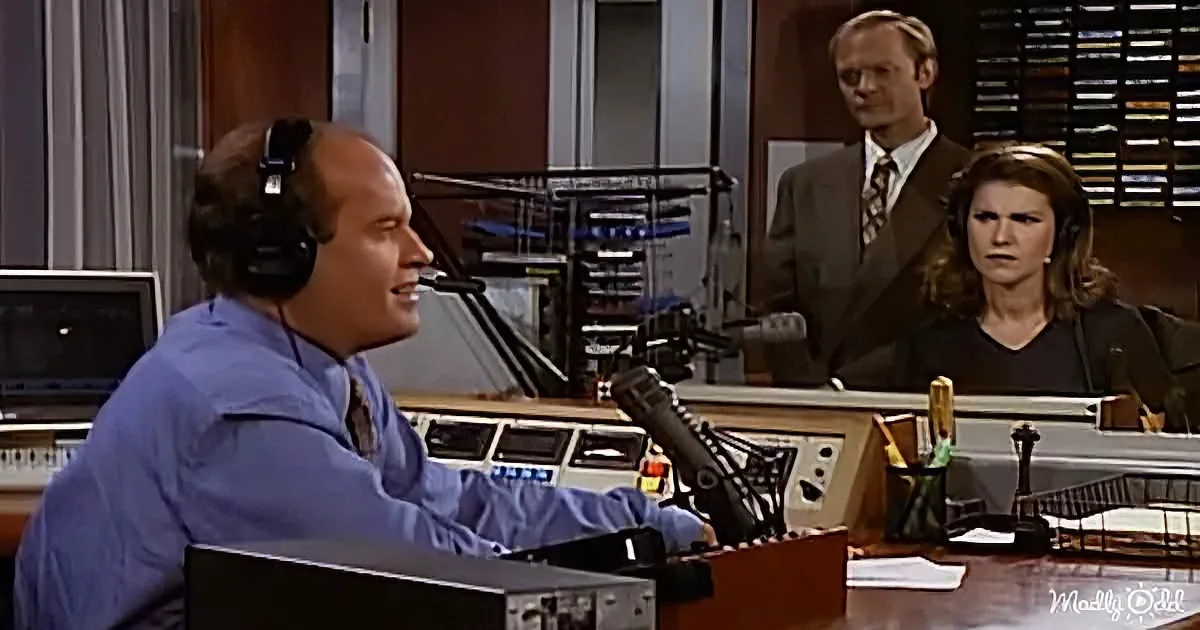 A man in a blue shirt and headphones sits at a desk with a microphone, speaking into it.