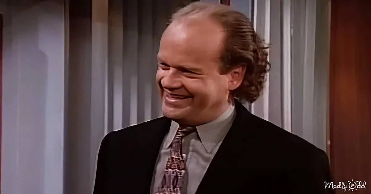 A man in a suit and tie smiles warmly, standing indoors with a neutral background.
