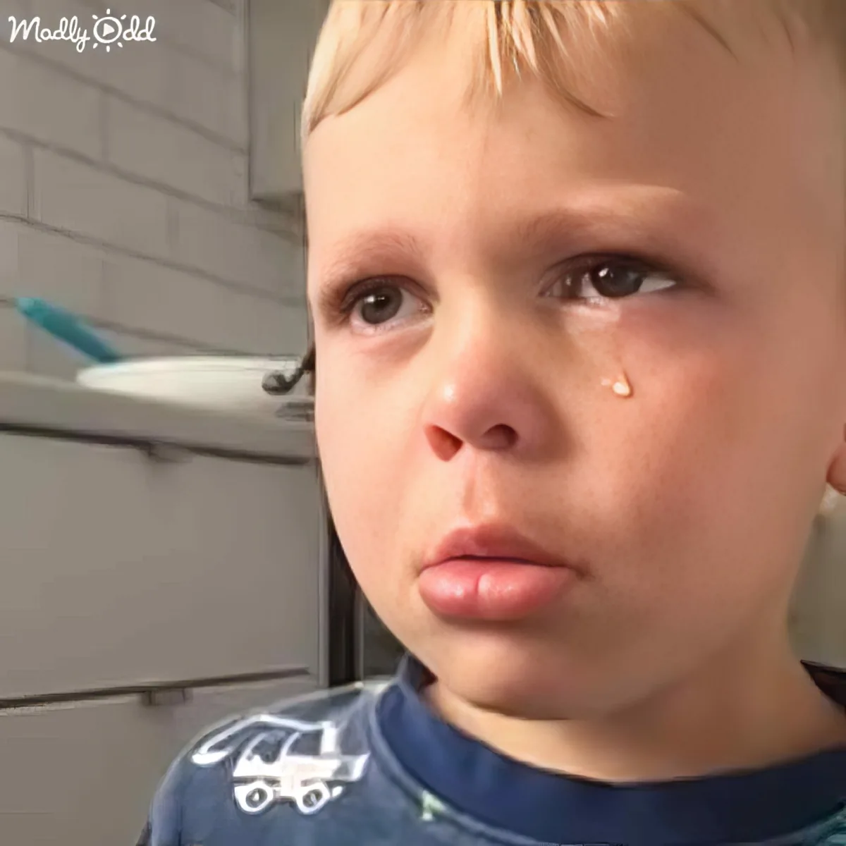 A young boy with blonde hair and a tear on his cheek looks sad, wearing a blue shirt with a car design.