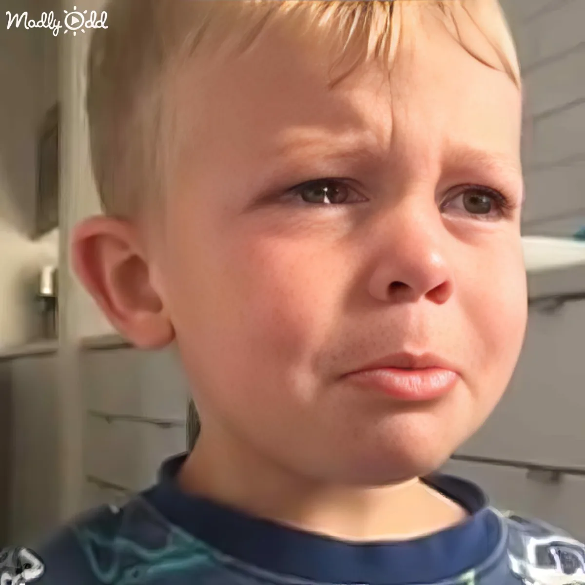 A young boy with short blonde hair and a blue shirt looks up with a concerned expression.