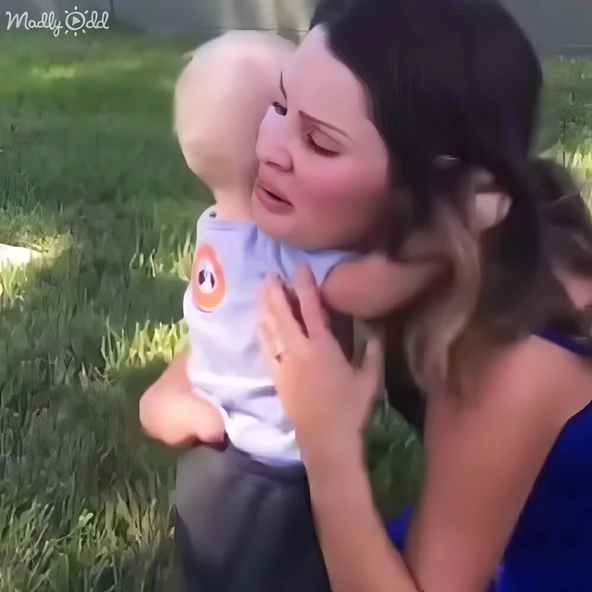 A woman holds a baby in a grassy area, both appear to be smiling and enjoying the moment.