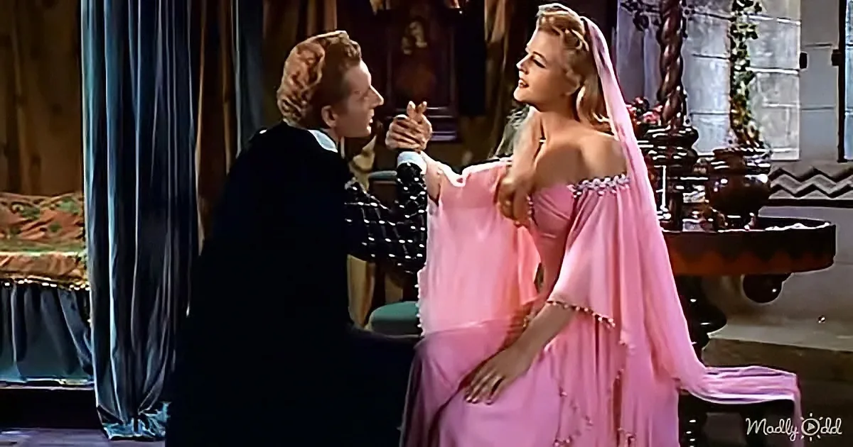 A man in a black suit holds hands with a woman in a pink gown, both in a richly decorated room.