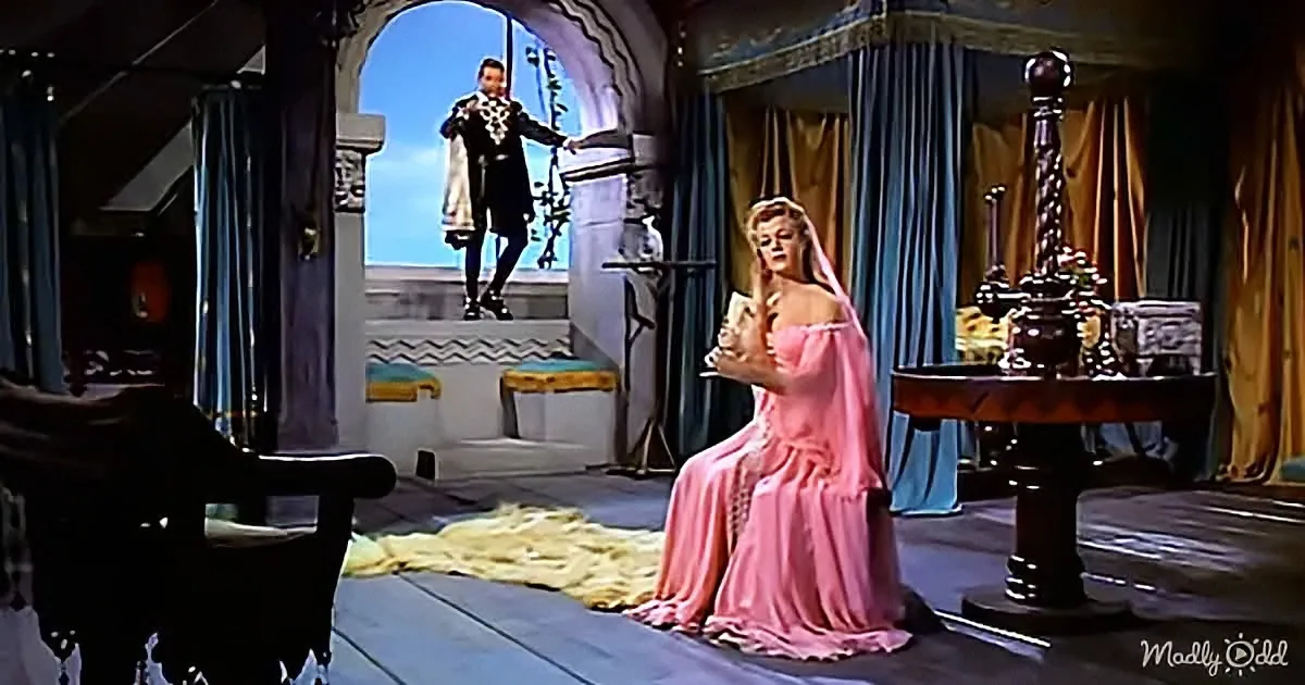 A woman in a pink dress sits on a stage, holding a book, while a man in a black and white outfit stands on a balcony above.