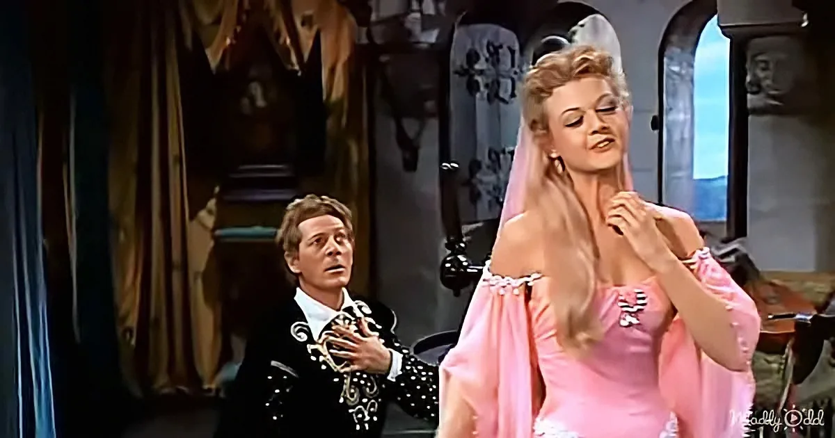 A woman in a pink dress stands beside a man in a black suit, both in a theatrical setting.