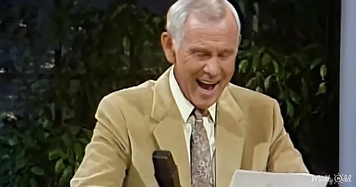A man in a beige suit and patterned tie is laughing, holding a microphone, with greenery in the background.