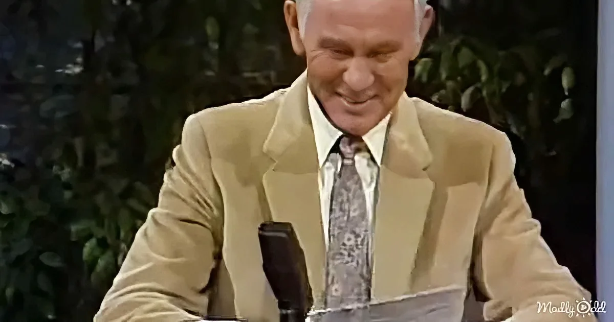 A man in a beige suit and patterned tie is smiling while seated at a desk with a microphone.