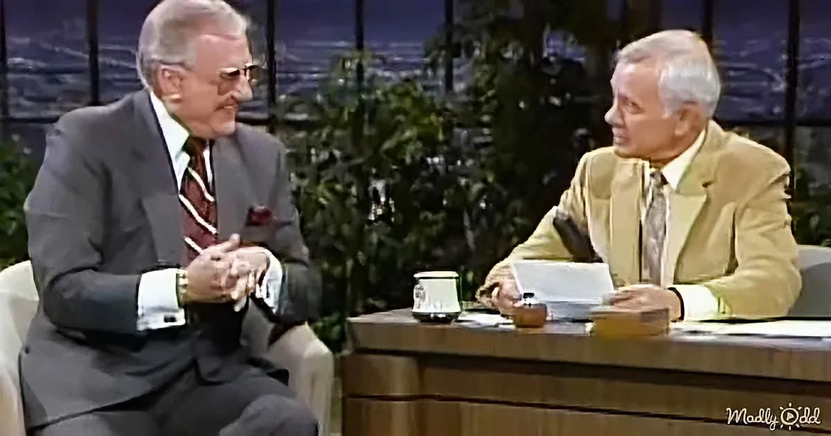 Two men in suits sit on a talk show set, one holding a mug, the other a script.