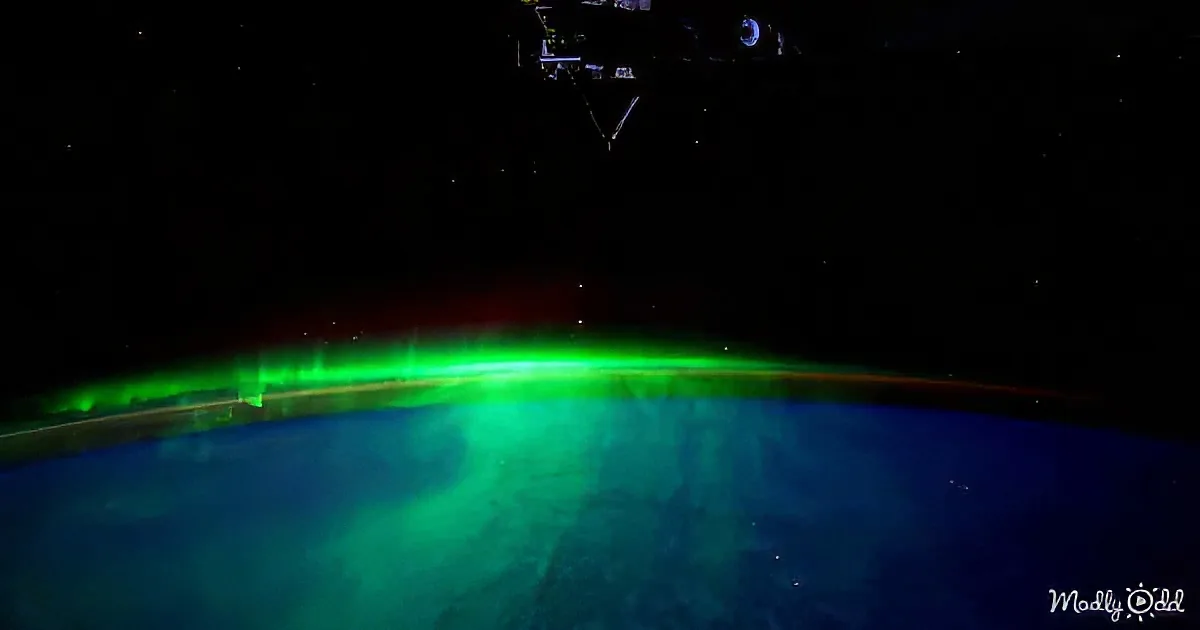 Aurora Borealis glows green over Earth's horizon, seen from space with stars and a satellite in view.
