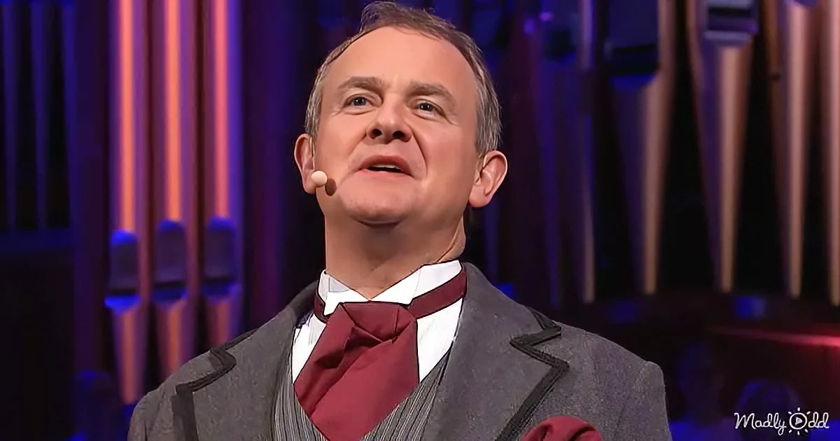 A man in a suit and bow tie stands on stage with a microphone, looking upwards.