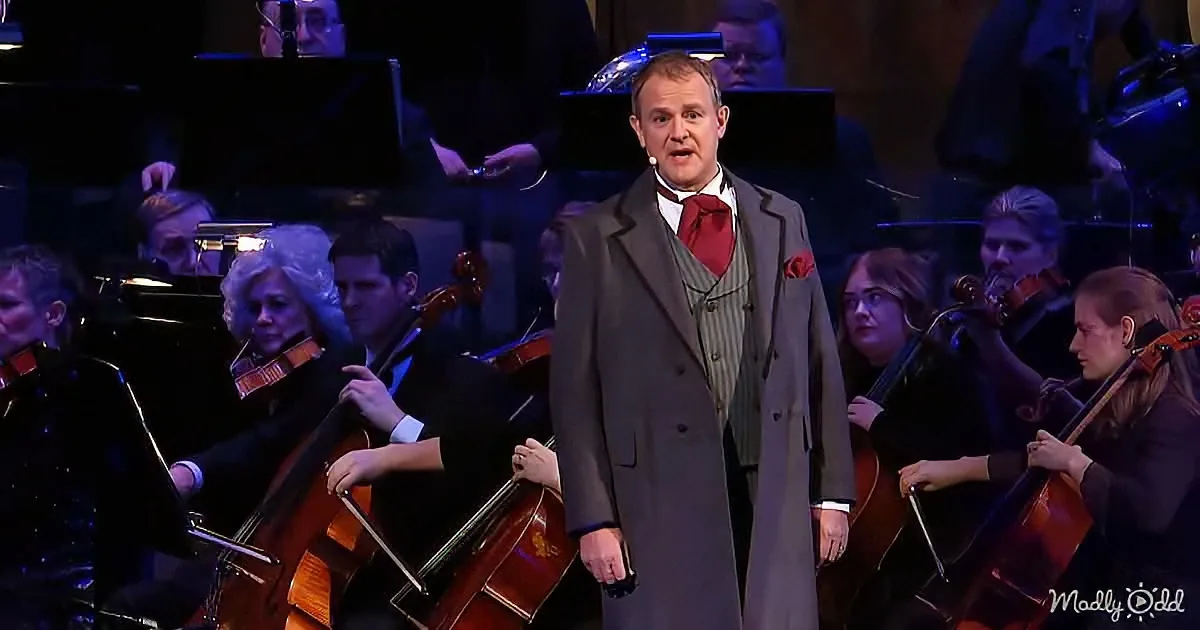 A man in a suit sings on stage with an orchestra playing in the background.