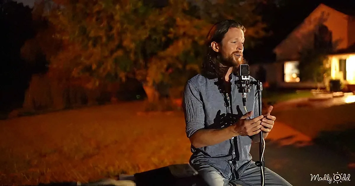 A man with long hair and beard speaks into a microphone at night, surrounded by trees and a lit building.