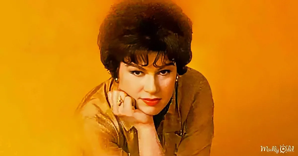 A woman with dark hair and red lipstick poses with her hand on her chin against an orange background.