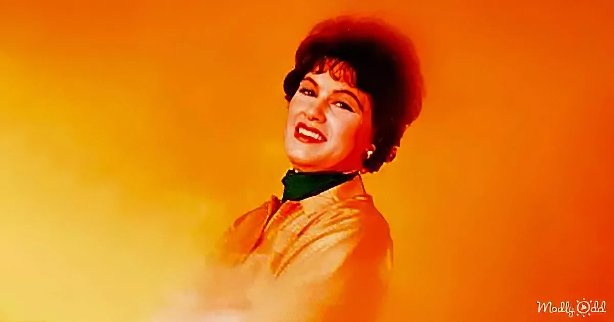 A woman with red hair, wearing a green scarf and a light brown jacket, smiles against a vibrant orange-yellow background.