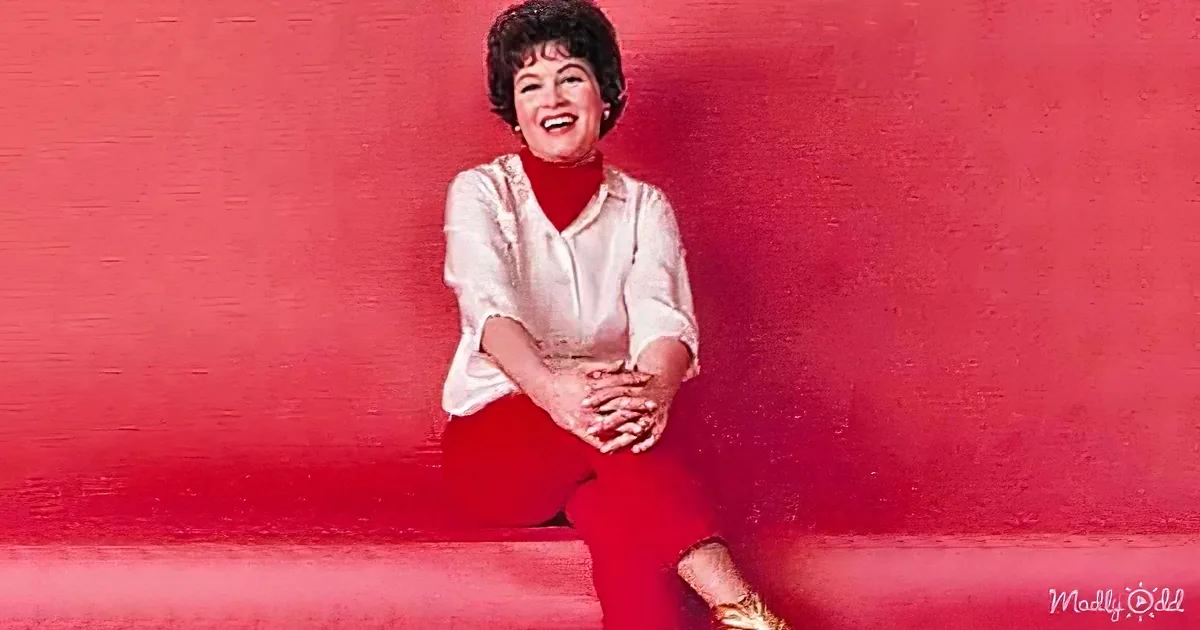 A woman in red pants and a white blouse sits on a red couch, smiling with hands clasped.