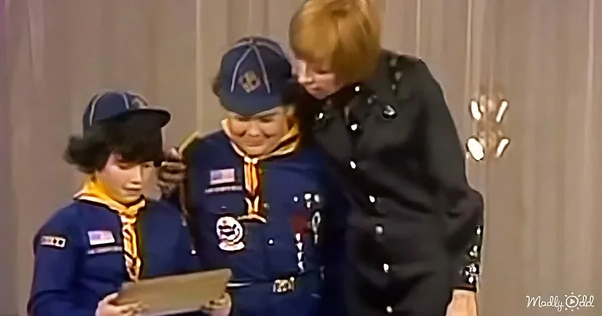 A young boy in a blue uniform and hat is being assisted by a woman in a black uniform, both looking at a book.