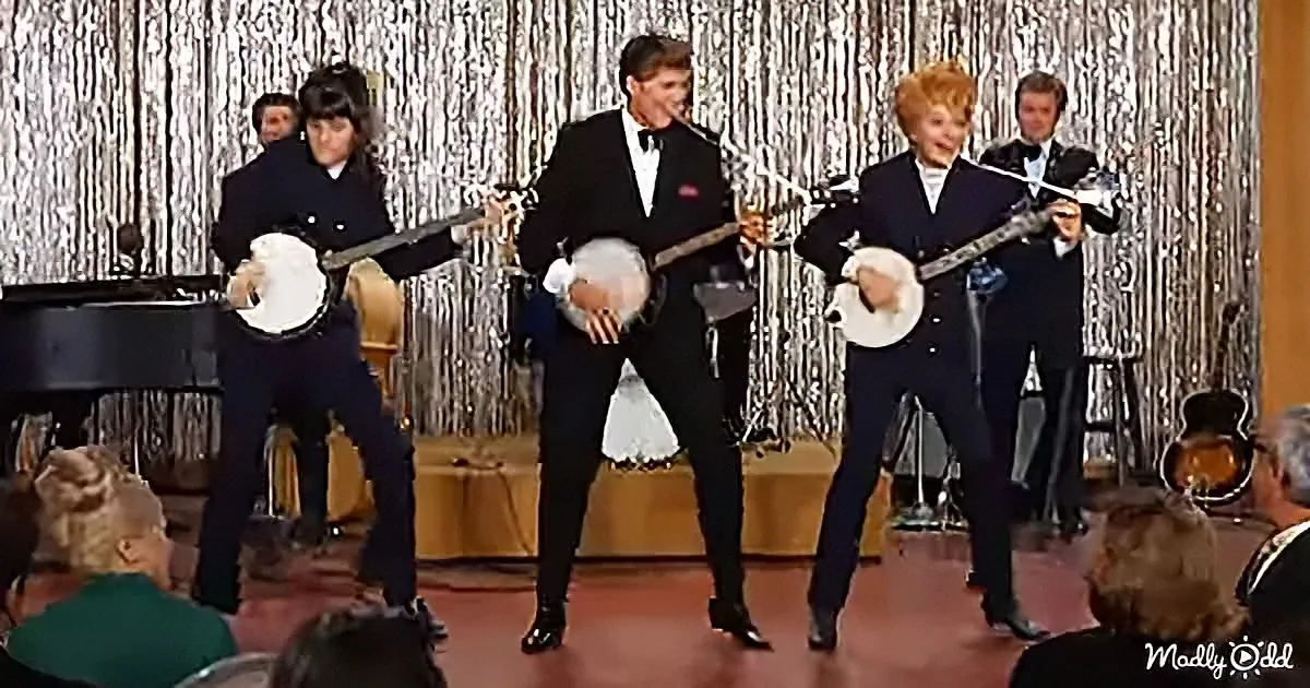 A group of musicians in suits play banjos on a stage with a silver curtain backdrop.