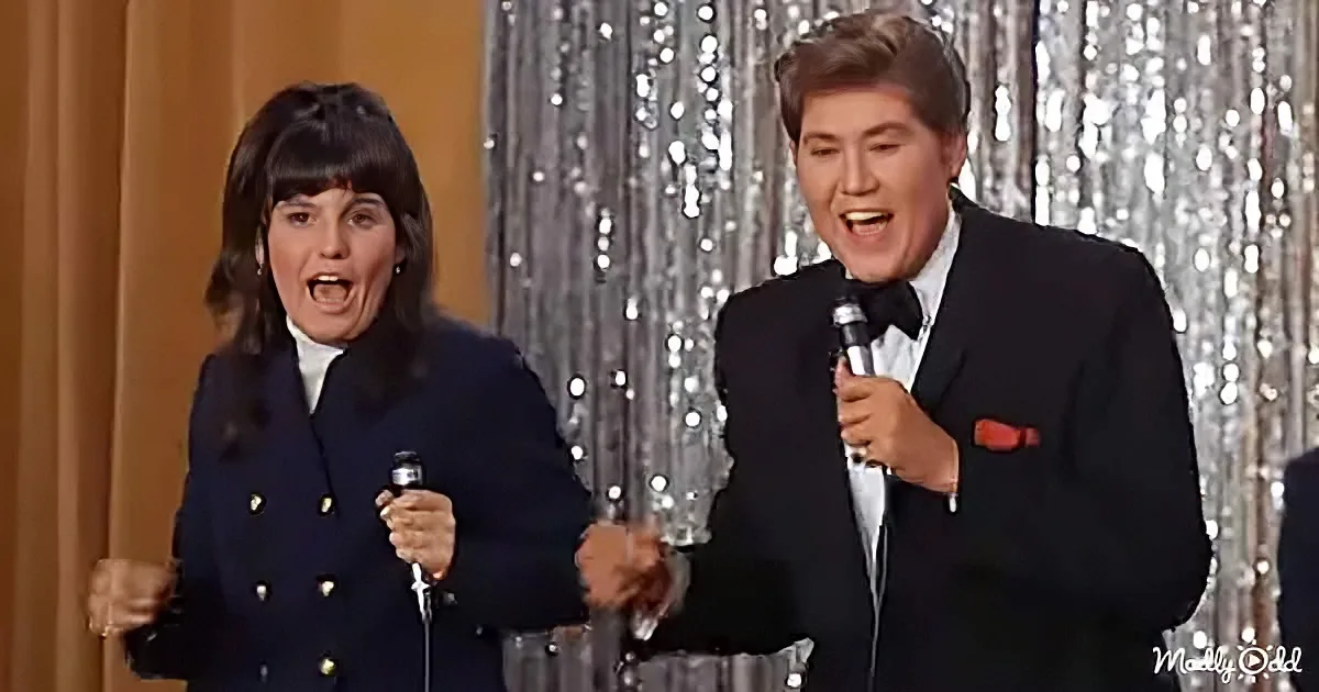 A man in a tuxedo and a woman in a navy coat sing into microphones on a stage with glittery curtains.