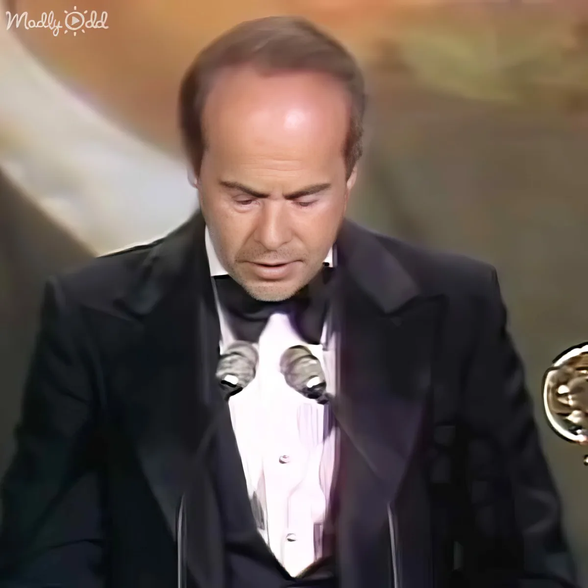 A man in a tuxedo stands at a podium, holding an award, looking down.