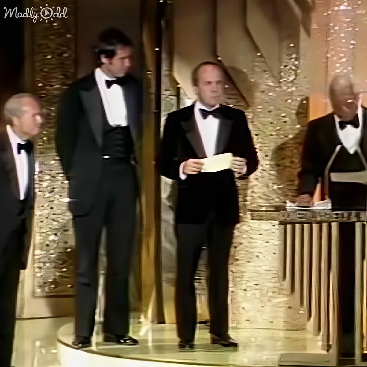 A man in a tuxedo stands on stage holding a card, surrounded by other formally dressed individuals.