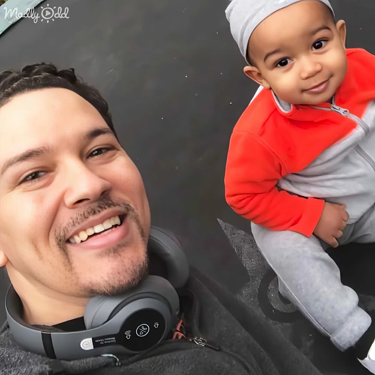 A smiling man with headphones around his neck holds a baby in a red and gray outfit.