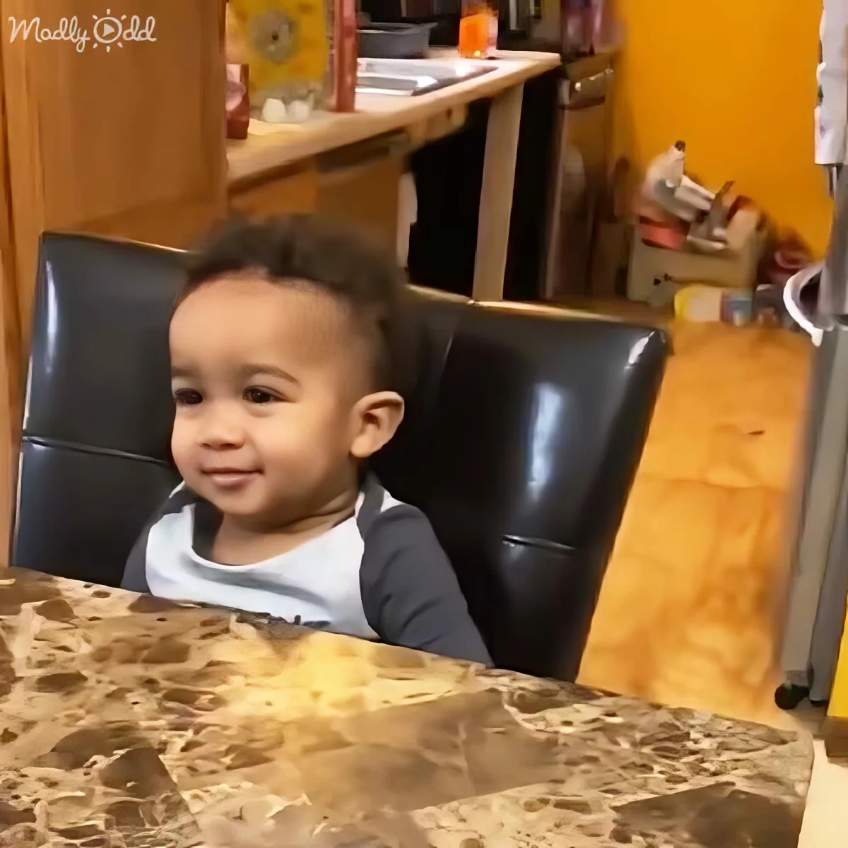 A smiling toddler sits in a black chair, wearing a white and blue shirt, in a room with a marble table and wooden floor.