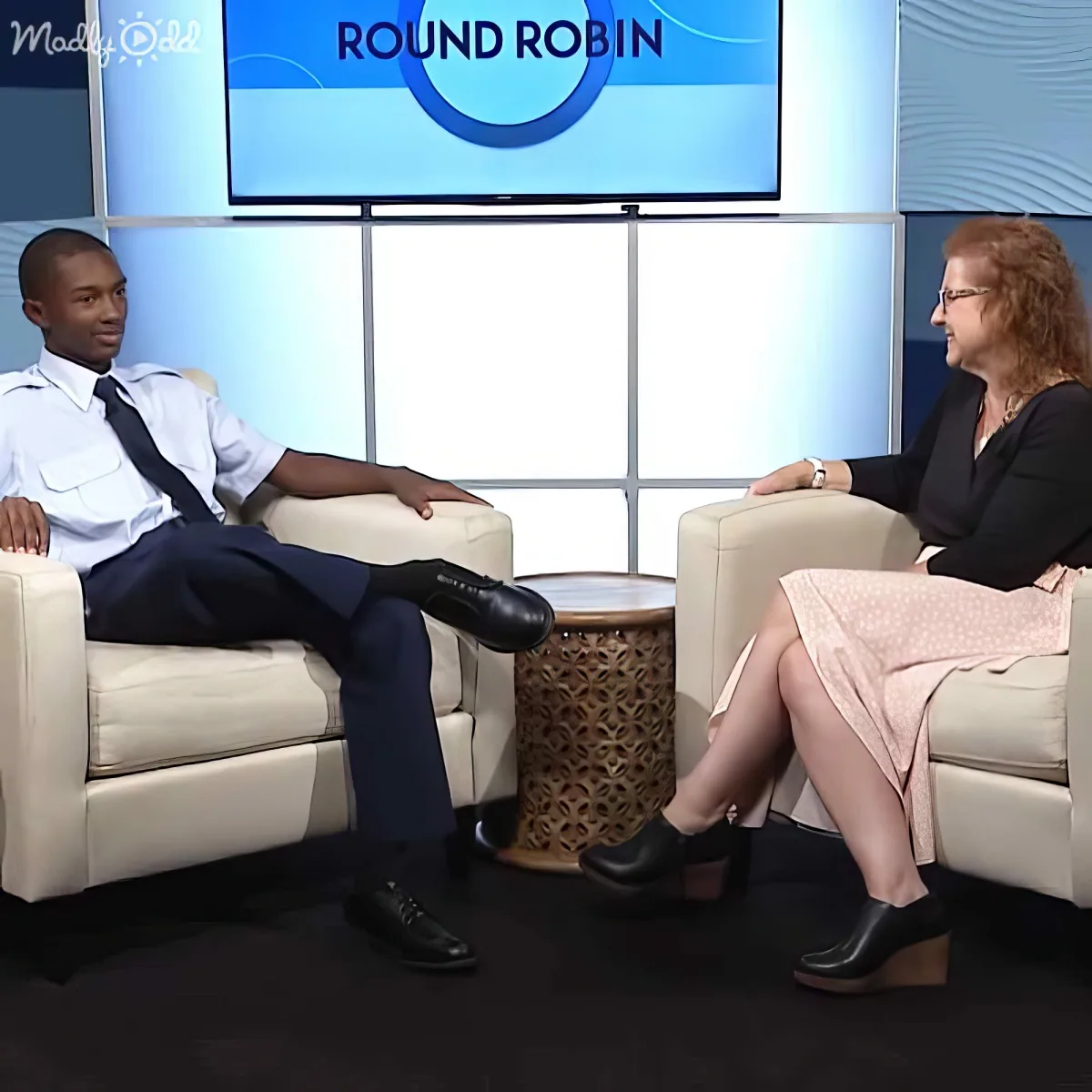 A man in a uniform and a woman in a dress sit in chairs, facing each other, on a set with a "Round Robin" sign.