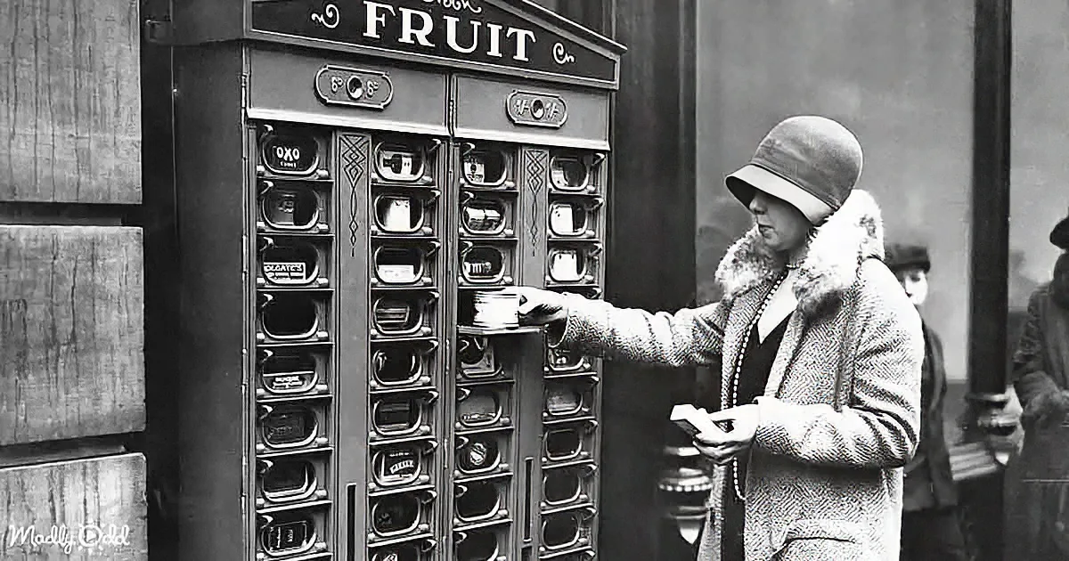 A woman in a fur coat and hat uses a vintage vending machine labeled "Fruit" to dispense items.