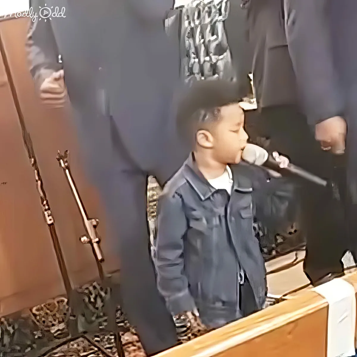 A young boy in a dark jacket sings into a microphone, surrounded by adults and a large inflatable figure.