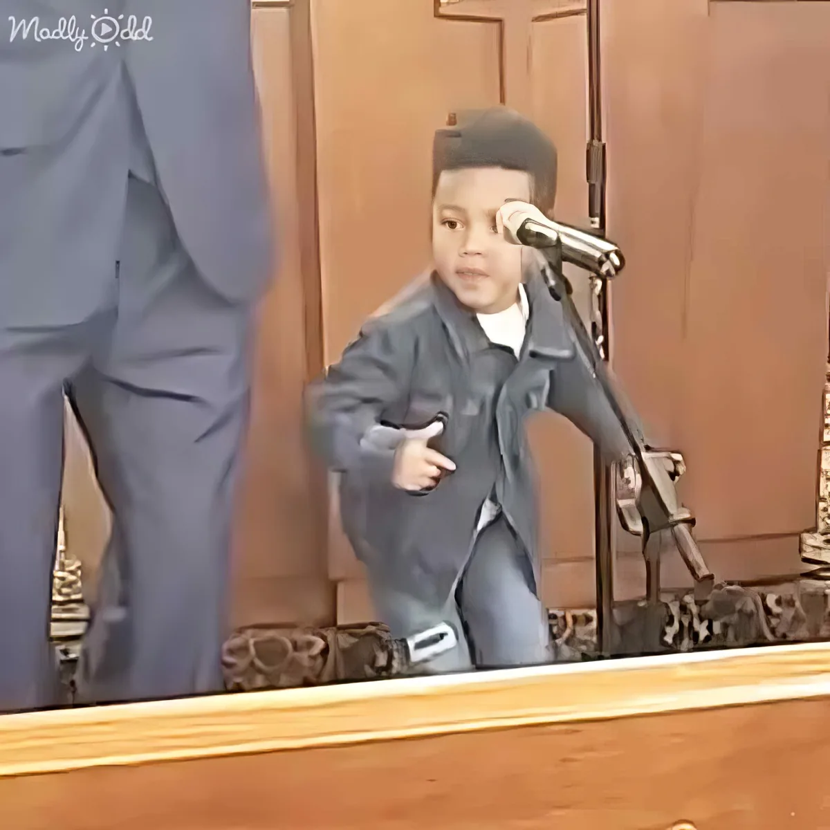 A young boy in a denim jacket stands at a podium with a microphone, gesturing with his hands.