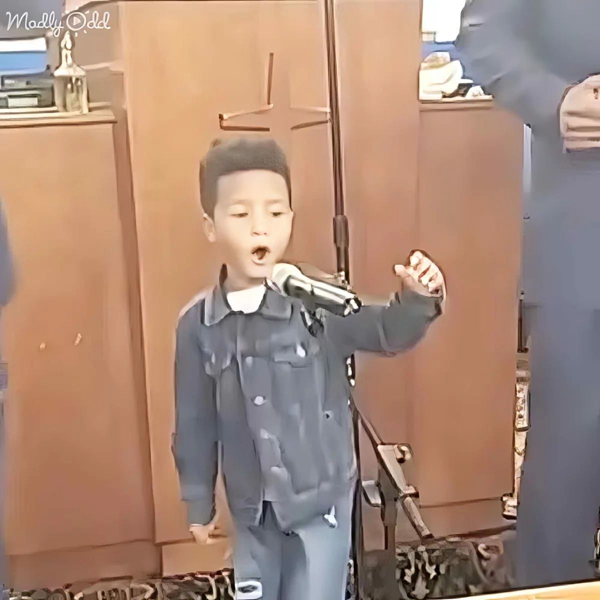 A young boy in a denim outfit sings into a microphone, raising his fist in a church setting.