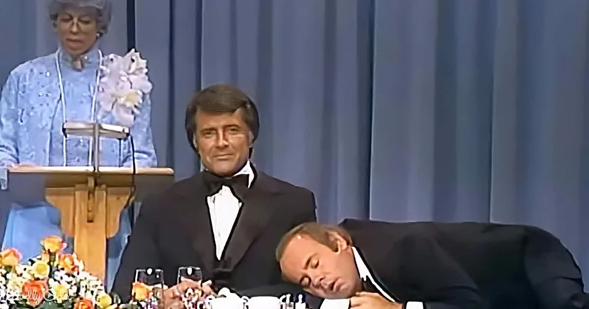 A man in a tuxedo leans forward at a table, appearing to sleep, while another man in a tuxedo sits upright behind him.