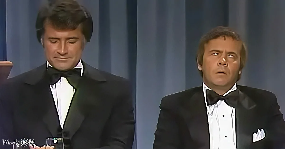 Two men in tuxedos, one holding a glass, both looking up, possibly at an award or presentation.