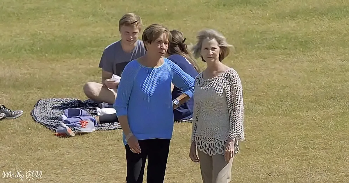 Two women in a grassy field, one in blue, the other in white, with a picnic blanket and people in the background.