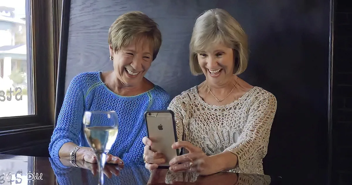 Two women are laughing, one holding a wine glass and the other a phone.
