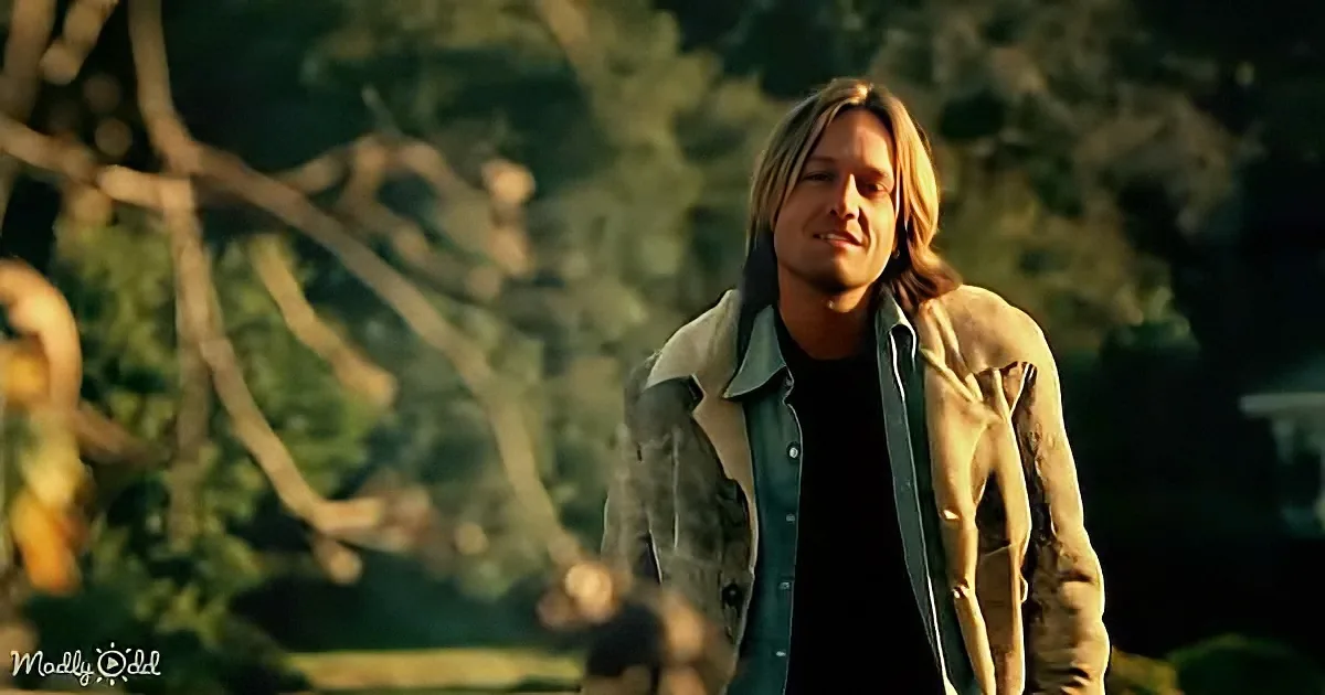A man with long hair, wearing a light brown jacket and a black shirt, stands outdoors with trees in the background.