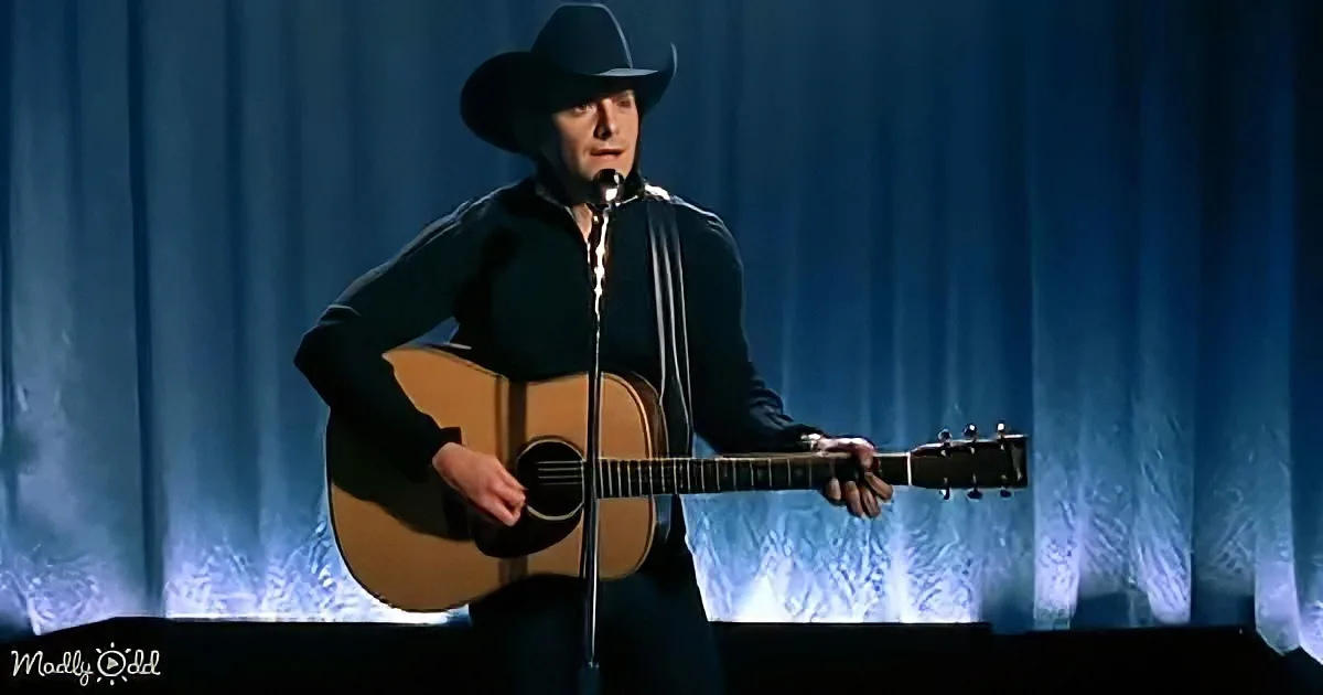 A man in a black cowboy hat and dark shirt plays an acoustic guitar on stage with a microphone.