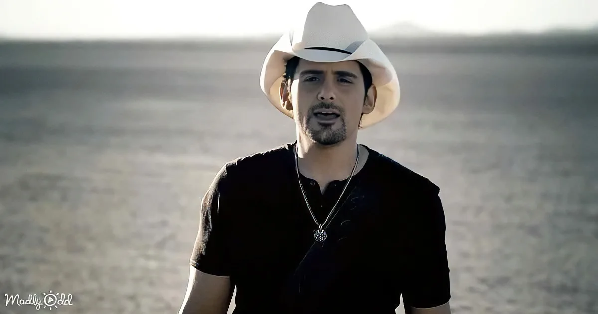 A man in a white cowboy hat and black shirt stands in a desert landscape, wearing a necklace.