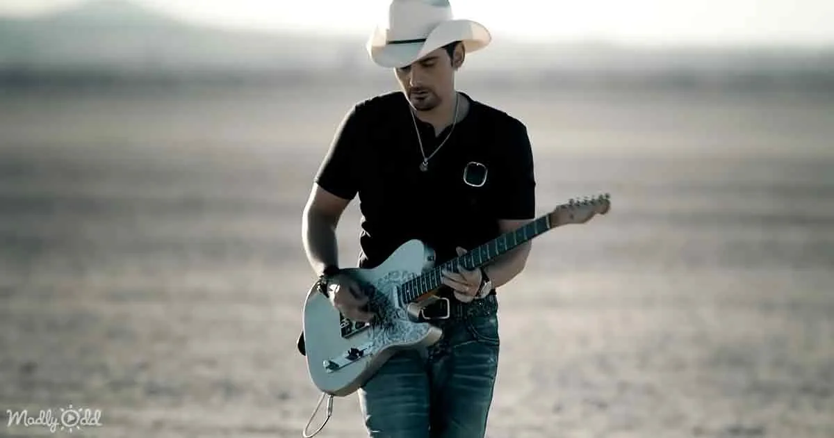 A man in a white hat plays a white electric guitar in a desert landscape.