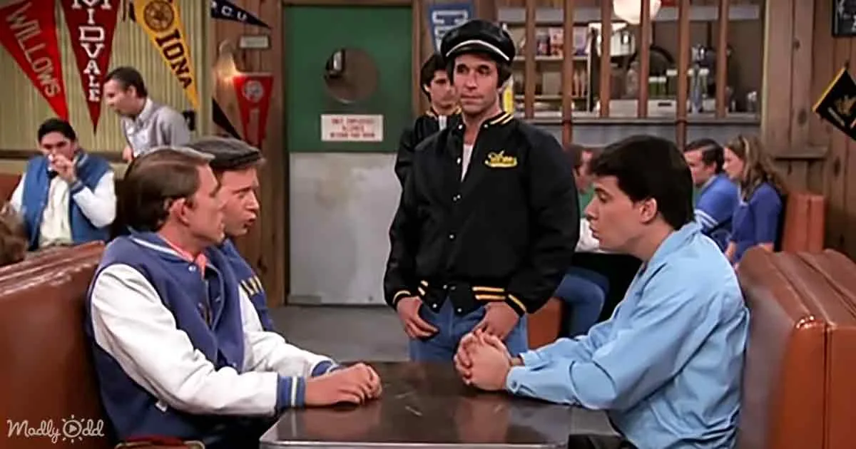 A man in a black uniform stands between two seated men in a diner, with other patrons and flags in the background.