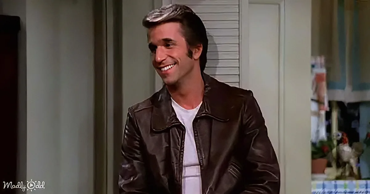 A man in a brown leather jacket smiles warmly, standing indoors near a door.