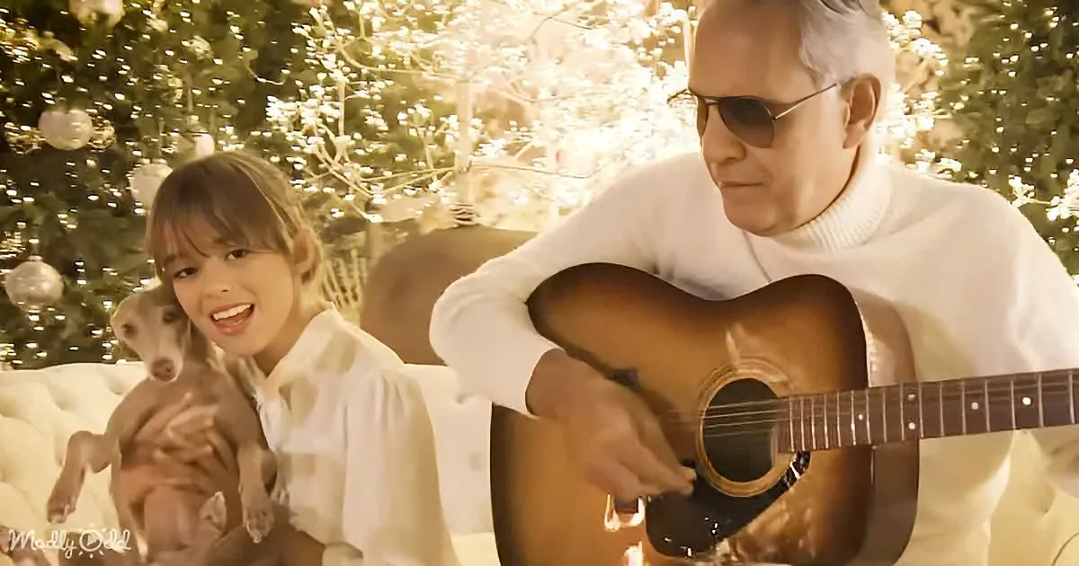 A man plays guitar while a woman holds a dog, both in front of a Christmas tree.