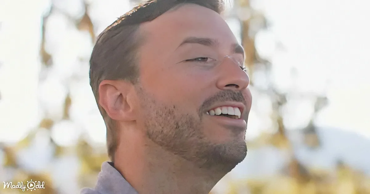 A man with short brown hair and a beard smiles, wearing a gray shirt, with a blurred background of yellow flowers.