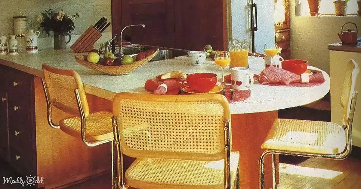A retro kitchen with a round table, yellow chairs, and a fruit bowl on the counter.